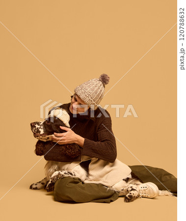 Young man in stylish warm winter clothes, chocolate sweater and knitted hat, hugging his purebred dog wearing sweater, against studio background Young man in stylish warm winter clothes, chocolate sweater and knitted hat, hugging his purebred dog wearing sweater, against studio background 120788632