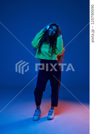 Full-length portrait of young Indian woman in casual clothes cheerfully listening to music in headphones against blue studio background in neon light Full-length portrait of young Indian woman in casual clothes cheerfully listening to music in headphones against blue studio background in neon light 120790090
