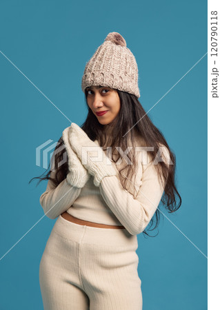 Portrait of young Indian woman wearing light outfit, knitted hat and gloves against blue studio background. Winter fashion 120790118