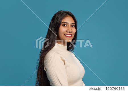 Side view portrait of young beautiful Indian woman with black hair, in light long sleeve, smiling against blue studio background 120790153