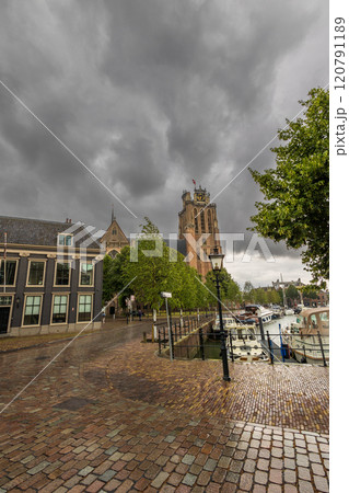 Historic European town with a clock tower under a dramatic cloudy sky. Cobblestone streets lead to a marina filled with boats. Trees and old architecture frame the peaceful scene. Dordrecht landscape Historic European town with a clock tower under a dramatic cloudy sky. Cobblestone streets lead to a marina filled with boats. Trees and old architecture frame the peaceful scene. Dordrecht landscape 120791189