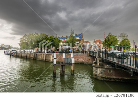 A canal scene with a historic drawbridge, lined with classic Dutch buildings and trees. Boats are docked under the cloudy sky, and a church tower is visible in the background. Dordrecht landscape. A canal scene with a historic drawbridge, lined with classic Dutch buildings and trees. Boats are docked under the cloudy sky, and a church tower is visible in the background. Dordrecht landscape. 120791190