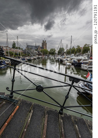 Serene harbor with moored boats under a dramatic cloudy sky. Quaint historic buildings and trees line the waterfront, reflecting in the calm green water, creating peaceful scene. Dordrecht landscape. Serene harbor with moored boats under a dramatic cloudy sky. Quaint historic buildings and trees line the waterfront, reflecting in the calm green water, creating peaceful scene. Dordrecht landscape. 120791191