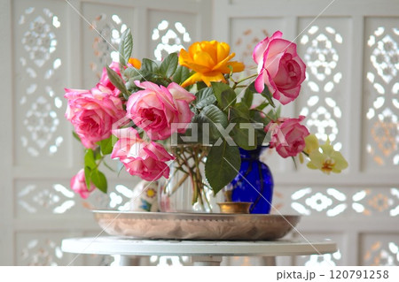 Pink and yellow roses in glass vase on metal tray on white round table. Background white wooden folding screen room divider. Floral arrangement as decoration for interior 120791258