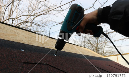 a man's hand with a screwdriver attaches soft tiles to the roof material of a small building, the final process of building a chicken coop under the roof 120791773