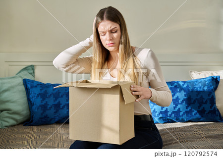 Upset sad confused young woman unpacking cardboard box parcel from online store 120792574