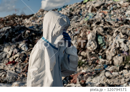 A worker dressed in protective clothing surveys a landfill, surrounded by piles of mixed waste. The bright sky contrasts with the grimy surroundings, highlighting environmental issues A worker dressed in protective clothing surveys a landfill, surrounded by piles of mixed waste. The bright sky contrasts with the grimy surroundings, highlighting environmental issues 120794571