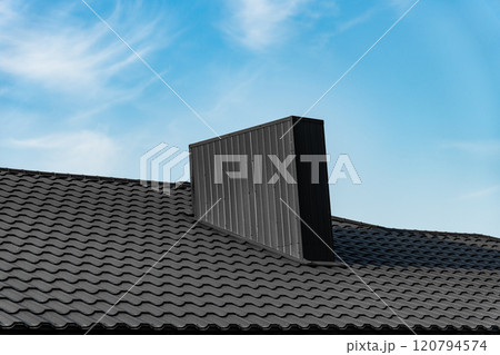 A modern, black chimney sits atop a black, tiled roof. The chimney is rectangular with vertical lines. The background is a blue sky with white clouds 120794574