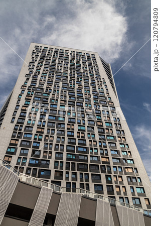 A modern high-rise building with a unique geometric facade, viewed from a low angle against a partly cloudy blue sky. The windows reflect sunlight, emphasizing its contemporary design. 120794809