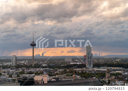 A cityscape at sunset with dramatic clouds, a prominent TV tower, modern skyscrapers, and a sprawling urban environment. The horizon features wind turbines and an industrial skyline. A cityscape at sunset with dramatic clouds, a prominent TV tower, modern skyscrapers, and a sprawling urban environment. The horizon features wind turbines and an industrial skyline. 120794815