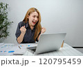 A woman is sitting at a desk with a laptop open and a smile on her face 120795459