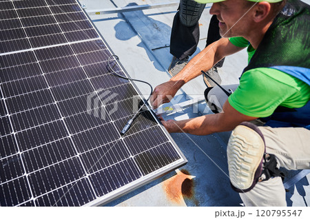 Workers building solar panel system on metal rooftop of house. Two men installers installing photovoltaic solar module outdoors. Alternative, green and renewable energy generation concept. Workers building solar panel system on metal rooftop of house. Two men installers installing photovoltaic solar module outdoors. Alternative, green and renewable energy generation concept. 120795547