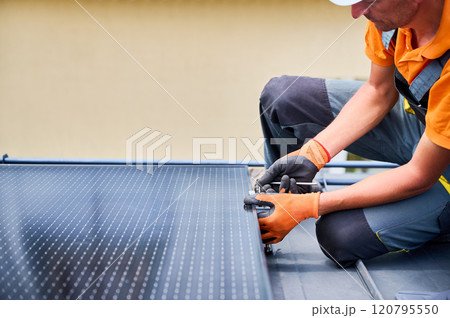 Worker building photovoltaic solar panel system on rooftop of house. Man engineer in gloves installing solar module with help of hex key outdoors. Alternative and renewable energy generation concept. 120795550