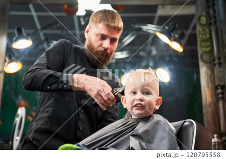 Cute little boy getting haircut. Blond curious kid first time at barbershop. Professional hairdresser making stylish haircut by shaver. 120795558
