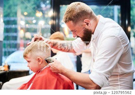 Barber using comb and shaver to cut hair. Professional hairdresser shaving little kid's nape. 120795562