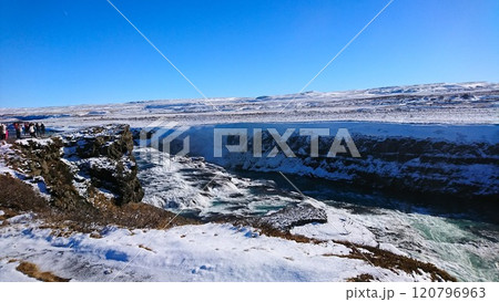 Gullfoss, Iceland - 3.24.2018: Tourists on the lookout viewing the breathtaking view of a roaring waterfall cutting through a rugged terrain towards icy water under a clear blue sky Gullfoss, Iceland - 3.24.2018: Tourists on the lookout viewing the breathtaking view of a roaring waterfall cutting through a rugged terrain towards icy water under a clear blue sky 120796963
