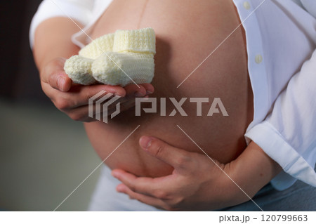 A pregnant woman gently cradles her baby bump while holding pair of knitted baby booties, symbolizing love and anticipation for new arrival A pregnant woman gently cradles her baby bump while holding pair of knitted baby booties, symbolizing love and anticipation for new arrival 120799663