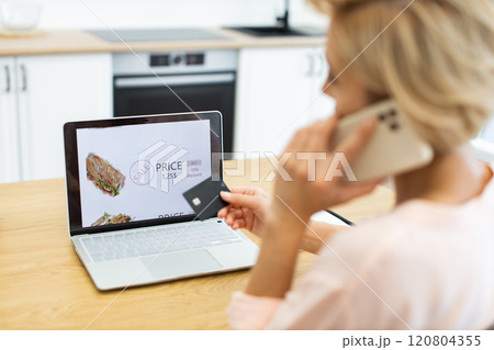 Caucasian woman orders food online using smartphone and laptop in kitchen. She browses meal options and pays with bank card. Image captures convenience of online shopping and digital payment. Caucasian woman orders food online using smartphone and laptop in kitchen. She browses meal options and pays with bank card. Image captures convenience of online shopping and digital payment. 120804355