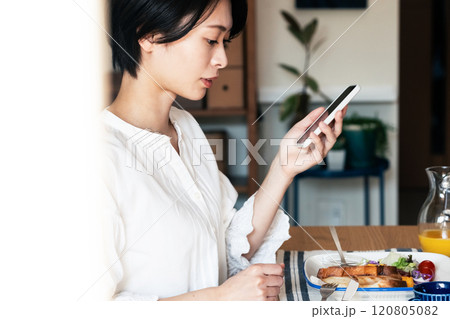スマホを見ながら朝食を食べる女性 スマホを見ながら朝食を食べる女性 120805082