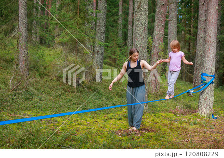 Teenage girl assists her sister balance on a slackline in the forest 120808229