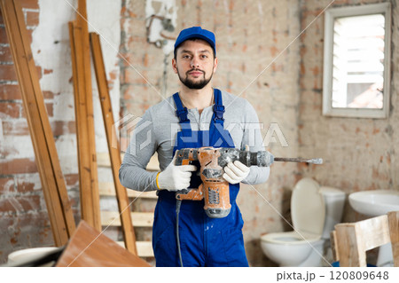 Young bearded builder holding pneumatic jackhammer in building under renovation 120809648