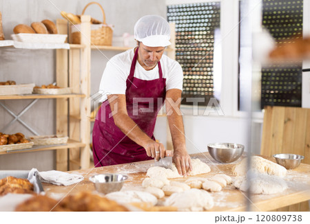 Caucasian man work in bakery as baker, cut dough into portions, forms pieces of dough to create buns Caucasian man work in bakery as baker, cut dough into portions, forms pieces of dough to create buns 120809738