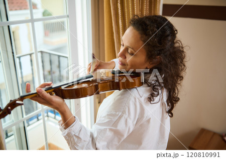 Woman playing violin near window with peaceful expression and natural light Woman playing violin near window with peaceful expression and natural light 120810991