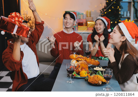 Group of young Asian man and women as friends having fun at a New Year's celebration, holding gift boxes standing by Christmas tree decoration, midnight countdown Party at home with holiday season. 120811864