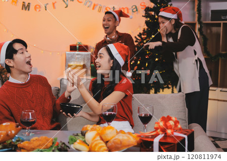 Group of young Asian man and women as friends having fun at a New Year's celebration, holding gift boxes standing by Christmas tree decoration, midnight countdown Party at home with holiday season. 120811974