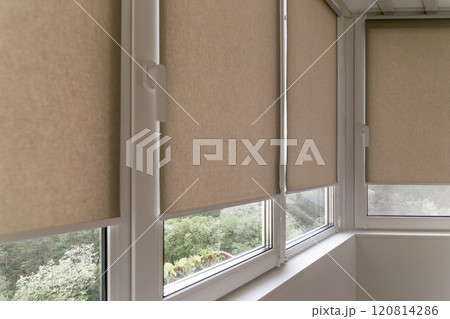 Beige roller blinds covering windows in a compact, bright balcony area. The sleek design offers privacy while allowing subtle natural light to filter through roller shades. Greenery outside. 120814286