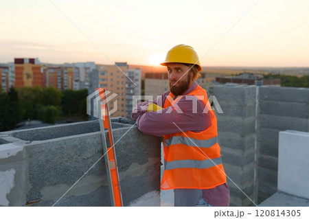 Construction worker wearing hardhat and safety vest leaning on concrete block at sunset Construction worker wearing hardhat and safety vest leaning on concrete block at sunset 120814305