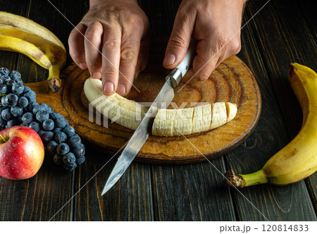 Hands skillfully slice a ripe banana on a wooden cutting board, surrounded by vibrant bananas, blueberries, and a crisp apple, showcasing fresh ingredients and culinary creativity Hands skillfully slice a ripe banana on a wooden cutting board, surrounded by vibrant bananas, blueberries, and a crisp apple, showcasing fresh ingredients and culinary creativity 120814833