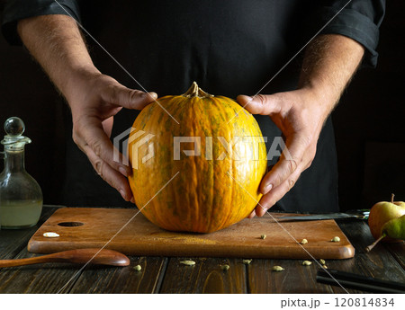 Hands gently cradle a bright orange pumpkin, ready for cooking. The rustic wooden table adds warmth, complemented by kitchen tools and a hint of green apples, evoking autumn creativity 120814834