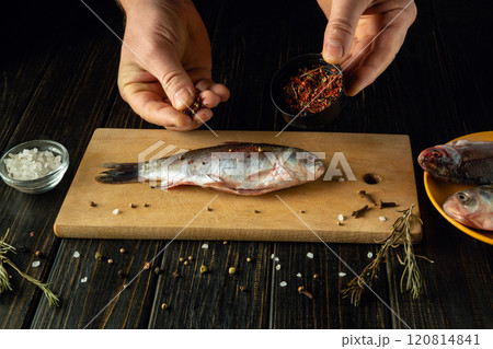 Hands expertly season a fresh fish with spices on a wooden cutting board. Surrounding ingredients include salt and herbs, creating an inviting atmosphere for culinary creativity. 120814841