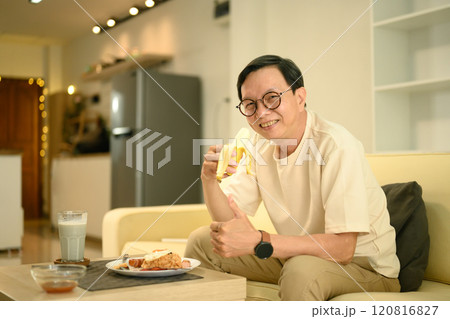 Cheerful middle aged man holding banana and enjoying his breakfast at cozy home Cheerful middle aged man holding banana and enjoying his breakfast at cozy home 120816827