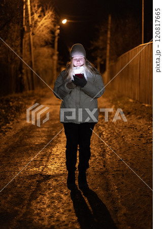 Young woman walking alone at night, illuminated by her smartphone on a quiet, dimly lit street with a rustic and peaceful atmosphere. 120817665