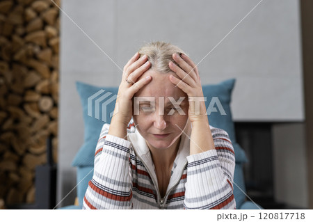 Middle-aged woman holding her head in her hands with closed eyes, appearing reflective and calm, seated indoors in a cozy atmosphere. 120817718