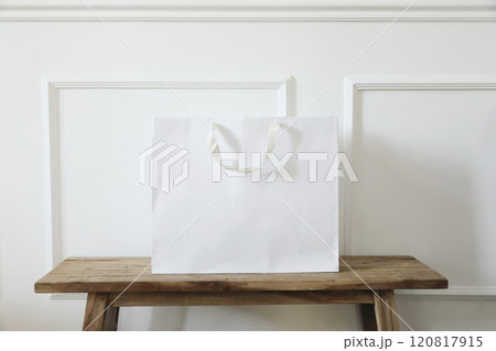 Clean simple white paper shopping bag mockup with handles on old wooden table, bench. White wall background with stucco decor. Empty gift bag for branding display, message. Retail business concept Clean simple white paper shopping bag mockup with handles on old wooden table, bench. White wall background with stucco decor. Empty gift bag for branding display, message. Retail business concept 120817915