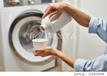 Woman pours liquid transparent laundry detergent or conditioner into plastic cap against washing machine. 120818846
