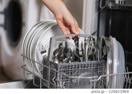 Female hand removing clean cutlery from a loaded dishwasher. Background of open wash machine  120823969