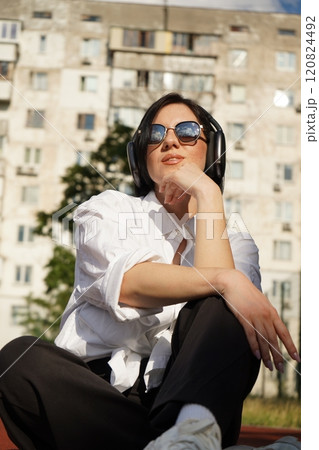 Woman in sunglasses and headphones sits outdoors on a track, enjoying a sunny day and her favorite tunes, vertical photo. 120824492