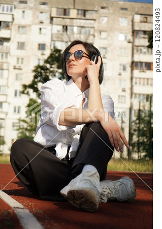 Woman in sunglasses and headphones sits outdoors on a track, enjoying a sunny day and her favorite tunes, vertical photo. 120824493