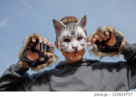 A playful child wearing a cat mask and furry gloves, posing with a cheerful expression against a clear blue sky. 120824757