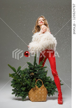 Woman in white fur coat and red leggings decorates Christmas tree in wicker basket with red ball, creative Christmas concept. Woman in white fur coat and red leggings decorates Christmas tree in wicker basket with red ball, creative Christmas concept. 120824787