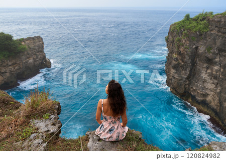 A beautiful woman in a pink dress sits on a cliff above the ocean on the island of Nusa Penida. Devil's Billabong an incredibly wonderful lagoon with splashes from the waves. 120824982
