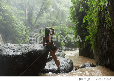 A young slender woman in a black swimsuit poses on a large rock in the water in the rain near a jungle waterfall on the popular island of Bali. 120825002