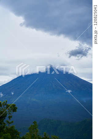 Panorama of Mount Agung and rice fields on the island of Bali. View of the mountain against a background of palm trees and a cornfield. Panorama of Agung volcano covered with clouds on a rainy day. Panorama of Mount Agung and rice fields on the island of Bali. View of the mountain against a background of palm trees and a cornfield. Panorama of Agung volcano covered with clouds on a rainy day. 120825041