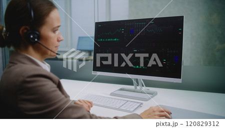 Female stock exchange trader talks on headset, monitors equity market and shares using computer 120829312