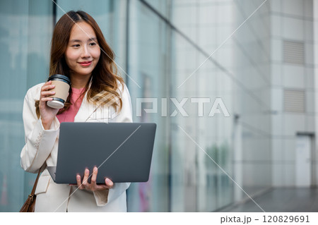 Confident businesswoman using computer laptop standing while drinking a cup of coffee takeaway at the city exterior. Beautiful smile female being ready to work. Business lifestyle concept. Confident businesswoman using computer laptop standing while drinking a cup of coffee takeaway at the city exterior. Beautiful smile female being ready to work. Business lifestyle concept. 120829691
