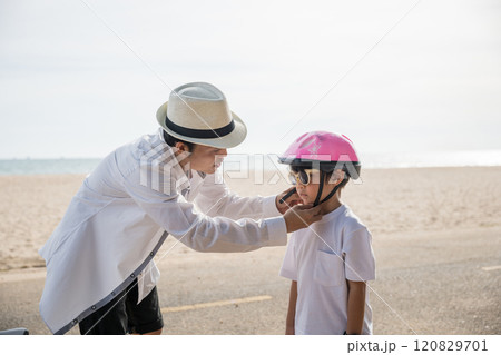 On a beach vacation a family experiences a heartwarming scene, the father in a safety helmet imparts the art of bicycle riding to his cheerful son creating an inspiring moment of learning and joy. On a beach vacation a family experiences a heartwarming scene, the father in a safety helmet imparts the art of bicycle riding to his cheerful son creating an inspiring moment of learning and joy. 120829701